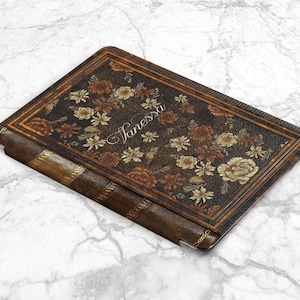 Pu&ograve; includere: Una custodia per tablet in pelle marrone con un design floreale e il nome "Janessa" in corsivo. La custodia ha l'aspetto di un libro vintage con dettagli dorati e un bordo decorativo.
