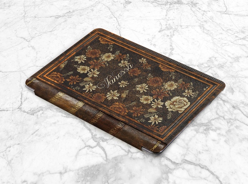 Pu&ograve; includere: Una custodia per tablet in pelle marrone con un design floreale e il nome "Janessa" in corsivo. La custodia ha l'aspetto di un libro vintage con dettagli dorati e un bordo decorativo.
