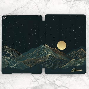 May include: A dark green tablet case featuring a mountain landscape design with gold-colored line art. The design includes a full moon and stars against a night sky. The name "Laura" is printed in gold script on the case.