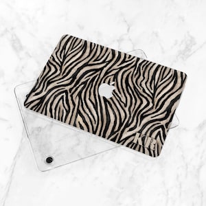 K&ouml;nnte beinhalten: Eine Laptop-H&uuml;lle mit einem Zebra-Print-Design in Schwarz und Beige. Die H&uuml;lle liegt auf einer transparenten Schutzh&uuml;lle. Das Apple-Logo ist sichtbar. Die Initialen "KRB" sind auf der H&uuml;lle aufgedruckt.