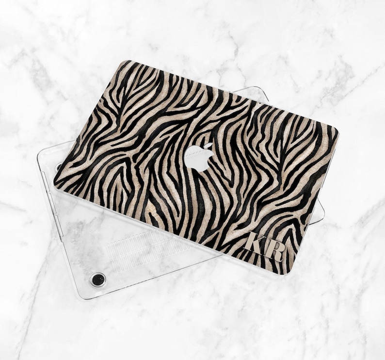 K&ouml;nnte beinhalten: Eine Laptop-H&uuml;lle mit einem Zebra-Print-Design in Schwarz und Beige. Die H&uuml;lle liegt auf einer transparenten Schutzh&uuml;lle. Das Apple-Logo ist sichtbar. Die Initialen "KRB" sind auf der H&uuml;lle aufgedruckt.