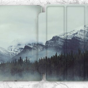 Puede incluir: Una funda de teléfono con un paisaje de montaña brumoso. Las montañas están cubiertas de nieve y los árboles se recortan contra el cielo.