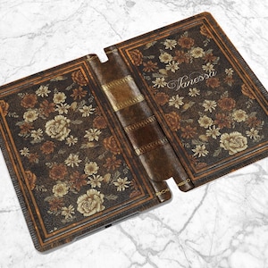 Pu&ograve; includere: Custodia per tablet aperta con un design a libro antico. La custodia &egrave; marrone con motivi floreali nei toni del marrone, beige e oro. Il nome "Vanessa" &egrave; scritto in corsivo sul lato destro.