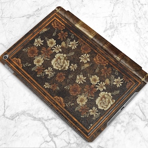 Pu&ograve; includere: Una custodia per iPad con un design di copertina di libro vintage. La custodia presenta una texture simile alla pelle marrone scuro con un motivo floreale nei toni del marrone e beige. I bordi hanno un bordo dorato.