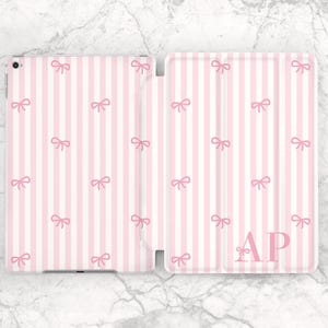 May include: Pink and white striped tablet case with a bow pattern. The case is open, revealing the interior. The initials "AP" are printed in pink on the front cover. The case is on a marble surface.