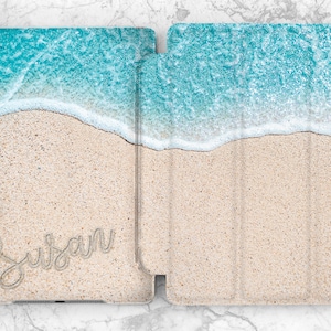 Könnte beinhalten: Eine weiße und blaue iPad-Hülle im Strandthema mit dem Namen "Susan", der in den Sand geschrieben ist.