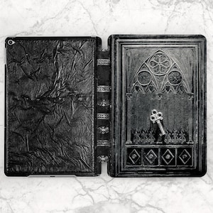 May include: A black tablet case featuring a textured, crinkled finish on the left side and an embossed gothic architectural design on the right side. A small, silver-toned metal key is placed centrally on the right panel of the case.
