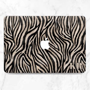 Puede incluir: Funda para portátil con estampado de cebra en negro y beige. El logotipo de Apple es visible, junto con las iniciales "KR". La funda tiene un acabado brillante. El diseño recuerda al estampado animal.