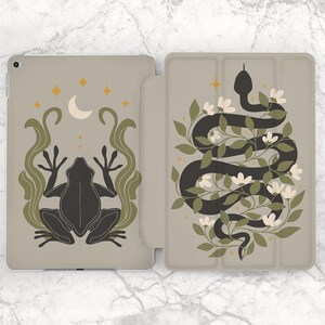 Puede incluir: Una funda de teléfono gris con una rana negra y una serpiente negra envuelta alrededor de enredaderas verdes con flores blancas sobre un fondo beige.