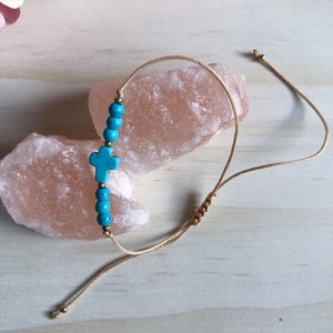 May include: A turquoise blue cross charm bracelet with gold beads and a beige cord. The bracelet is adjustable with a sliding knot.