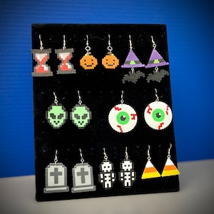 May include: A display of Halloween-themed earrings. Designs include an hourglass, jack-o'-lanterns, witch hats with bats, green alien heads, eyeballs, tombstones, skeletons, and candy corn. The earrings are made of colorful beads and have silver hooks.