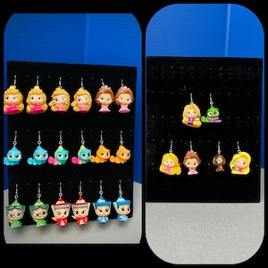 May include: A display of 18 pairs of dangle earrings featuring Disney characters. The earrings are displayed on a black background with a white grid pattern. The characters include princesses, a chameleon, and a squirrel.
