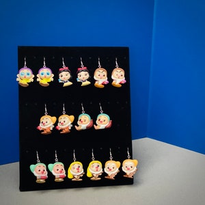 May include: A display of handmade character earrings. The earrings feature various cartoon characters in vibrant colors, including yellow, red, and brown. The earrings are displayed on a black velvet surface against a blue backdrop.