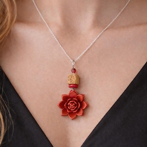 Może przedstawiać: Srebrny naszyjnik z czerwoną zawieszką w kształcie kwiatu. Zawieszka ma element przypominający korek i czerwone koraliki. Naszyjnik jest noszony na czarnym topie.