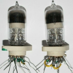 May include: Two vintage vacuum tubes with glass enclosures and multiple metal pins. The tubes are mounted on white ceramic bases with wires attached to the pins.