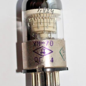 May include: A silver vacuum tube with a glass envelope and a black cap. The tube has a white ceramic base with a grid of metal wires. The tube is labeled "XH-70" and has a purple diamond with a letter "D" inside. The tube also has a green diamond with the number "4" inside.