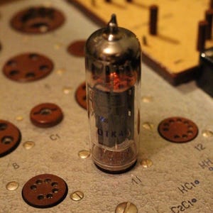 Puede incluir: Primer plano de un tubo de vacío de vidrio con una base de metal. El tubo está etiquetado como "OTKA" y tiene un brillo rojo en su interior. El tubo está sentado en una placa de circuito con varios componentes y etiquetas, incluyendo "C1", "10", "11", "12", "HC10" y "C2C10".