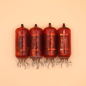 Può includere: Quattro tubi nixie cilindrici in vetro rosso disposti in fila. Ogni tubo presenta una struttura interna metallica con filamenti visibili e un gruppo di sottili pin metallici che sporgono dalla base per il collegamento elettrico.