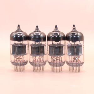 May include: Four vintage vacuum tubes with clear glass bulbs and dark metal tops. Each tube has internal components and a series of pins at the base. The tubes are arranged in a row against a light background.