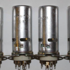 May include: Four vintage vacuum tubes with silver metal casings, each with a circular base and multiple pins. The tubes have text markings and a small rectangular window. The bases are white with a metal ring.