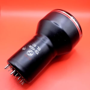 Pode incluir: Um componente eletrônico preto com um corpo cilíndrico e um topo alargado, com uma borda prateada. O componente tem uma série de pinos na base e o texto "06 79", "ФЗУ-82" e um logotipo. O fundo é vermelho liso.