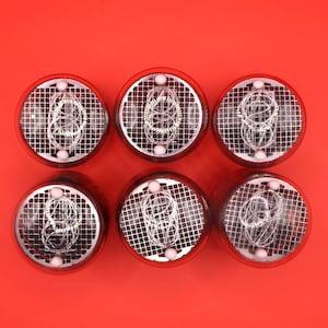 Op de afbeelding: Zes zilverkleurige Nixie-buizen met een rasterpatroon en gloeiende rode segmenten. De buizen zijn in een rij op een rode achtergrond gerangschikt.