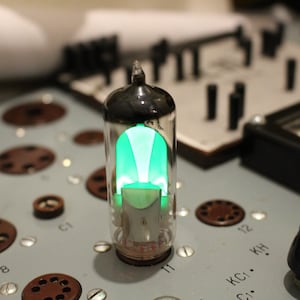 Puede incluir: Un tubo de vacío de vidrio transparente con un brillo verde en su interior. El tubo está sentado en una placa de circuito con varios botones y diales.