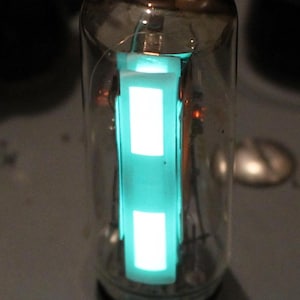 May include: A clear glass vacuum tube with a glowing green filament inside. The filament is shaped like a rectangle with two sections.