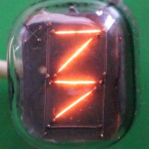 Może przedstawiać: Vintage'owa lampa Nixie wyświetlająca cyfrę "7" z jasnym pomarańczowym blaskiem. Lampa jest zamknięta w przezroczystej szklanej bańce z metalową podstawą.