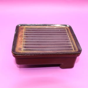 Può includere: Un vassoio rettangolare in ceramica marrone scuro caratterizzato da una superficie piana rialzata con una serie di scanalature orizzontali parallele. Il vassoio presenta una finitura lucida marrone scuro con sottili riflessi marrone chiaro sui bordi.