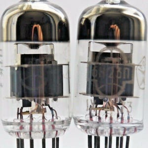 May include: Two vintage vacuum tubes with clear glass and silver metal tops. The tubes have a black cylindrical component inside and a label that reads "6SN7GT". The tubes have nine pins on the bottom.