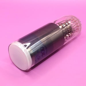 Puede incluir: Un tubo de vacío vintage con una carcasa de vidrio transparente, un componente cilíndrico oscuro y una base blanca. El tubo tiene componentes internos y marcas. El fondo es de color rosa sólido.