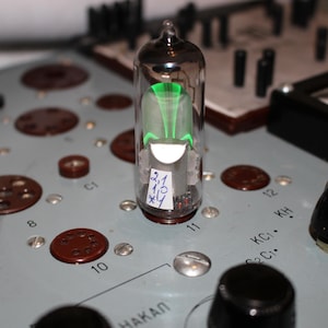 Puede incluir: Un tubo de vidrio transparente con un brillo verde en su interior. El tubo está etiquetado con "2,1,10, xly". El tubo está sobre una superficie gris con botones negros y marrones.