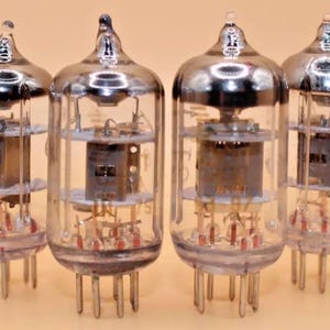 May include: Four vintage glass vacuum tubes with metal caps and clear glass bodies. The tubes have multiple internal components and are arranged in a row on a light brown surface.