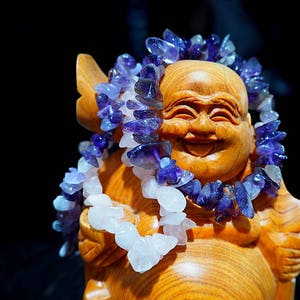 Peut inclure: Une sculpture en bois d'un Bouddha souriant portant un collier de pierres précieuses violettes et blanches.