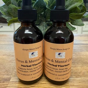 May include: Two brown glass bottles of herbal tincture with black droppers. The labels read "Sleepy Moon Soap Co. Focus & Mental Clarity Herbal Tincture." Each bottle contains 2 fl oz. The background features green foliage.