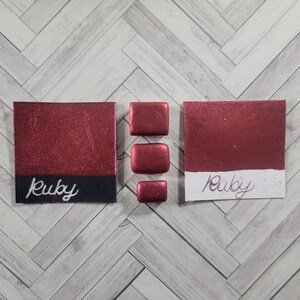 May include: A close-up of a red watercolor paint pan with the word "Ruby" written on it. The pan is surrounded by four smaller pans of the same color.