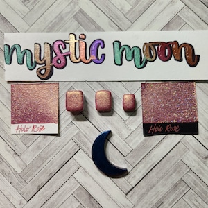 May include: A set of three small, square, metallic gold samples of "Holo Rose" nail polish. The samples are arranged in a row on a wooden surface. A larger sample of the same polish is shown in the background, along with a blue crescent moon shape.
