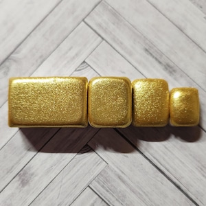 Könnte beinhalten: Vier quadratische Boxen in Goldtönen, in verschiedenen Größen, in einer Reihe vom größten zum kleinsten angeordnet. Die Boxen haben eine glitzernde Oberfläche.