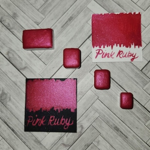 May include: Four watercolor paint pans in a pink ruby color. The pans are arranged in a row on a white surface. The paint is labeled "Pink Ruby".