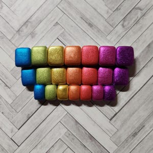 Könnte beinhalten: Eine Reihe kleiner, quadratischer, glitzernder Fliesen in Regenbogenfarben. Die Fliesen sind in einer Reihe angeordnet, wobei die Farben von Blau nach Lila übergehen.