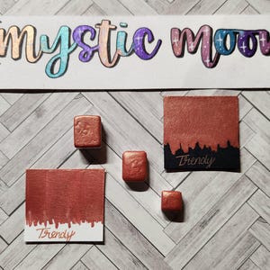 Pode incluir: Quatro pequenas bandejas de tinta de cor cobre com a palavra "Trendy" escrita em duas delas. As bandejas de tinta estão sobre uma superfície branca com um padrão de grão de madeira claro.