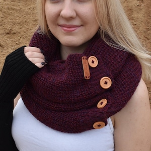 Puede incluir: Primer plano de una mujer con una bufanda infinita color burdeos con botones de madera. La bufanda está tejida y tiene un aspecto texturizado. La mujer lleva una camisa blanca y guantes sin dedos negros.
