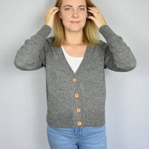 Puede incluir: Un cárdigan gris con botones de madera. El cárdigan está tejido y tiene cuello en V.