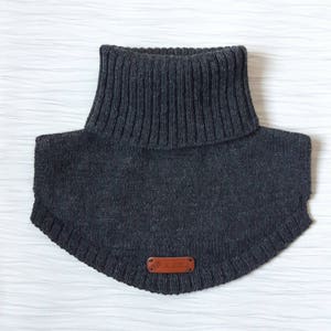 May include: A dark charcoal gray knitted neck warmer featuring a ribbed turtleneck collar and a curved hemline. A small, rectangular brown leather-like tag is stitched onto the lower front center, displaying the embossed text Hand Made.