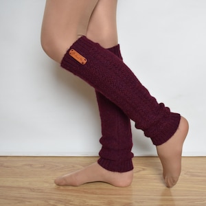May include: A pair of burgundy knitted leg warmers with a brown leather tag. The leg warmers are worn on a person's legs.