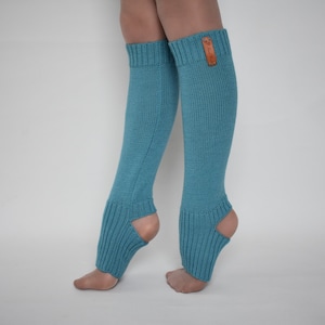 May include: A pair of teal blue knit leg warmers featuring a ribbed texture, an open heel design, and a small rectangular brown leather-like brand tag attached to the upper cuff of the right leg warmer, extending from the calf to the ankle.