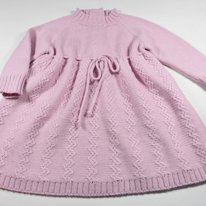 May include: A light pink long-sleeve knit dress featuring a ribbed mock neck, a drawstring waist with matching ties, and a vertical zigzag textured pattern throughout the skirt. A small brown rectangular tag is attached to the bottom right hem.