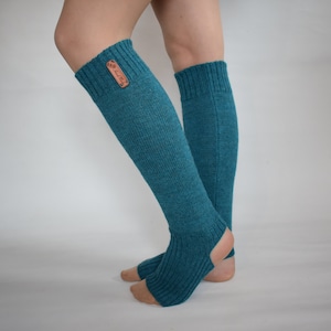 May include: A pair of teal, knee-high, open-heel leg warmers featuring a ribbed knit texture and a small, rectangular brown leather-like tag attached to the upper cuff. The design includes an opening for the heel and toes, exposing the skin.