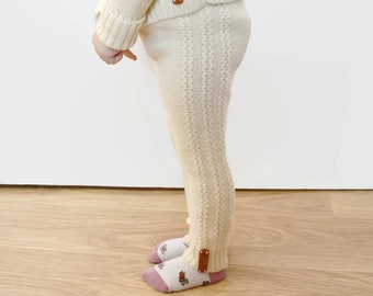 May include: Cream-colored knit leggings featuring a vertical cable-knit pattern along the outer leg. The leggings have a ribbed waistband and ribbed cuffs at the ankles, each accented with a small, rectangular brown leather-like brand tag.
